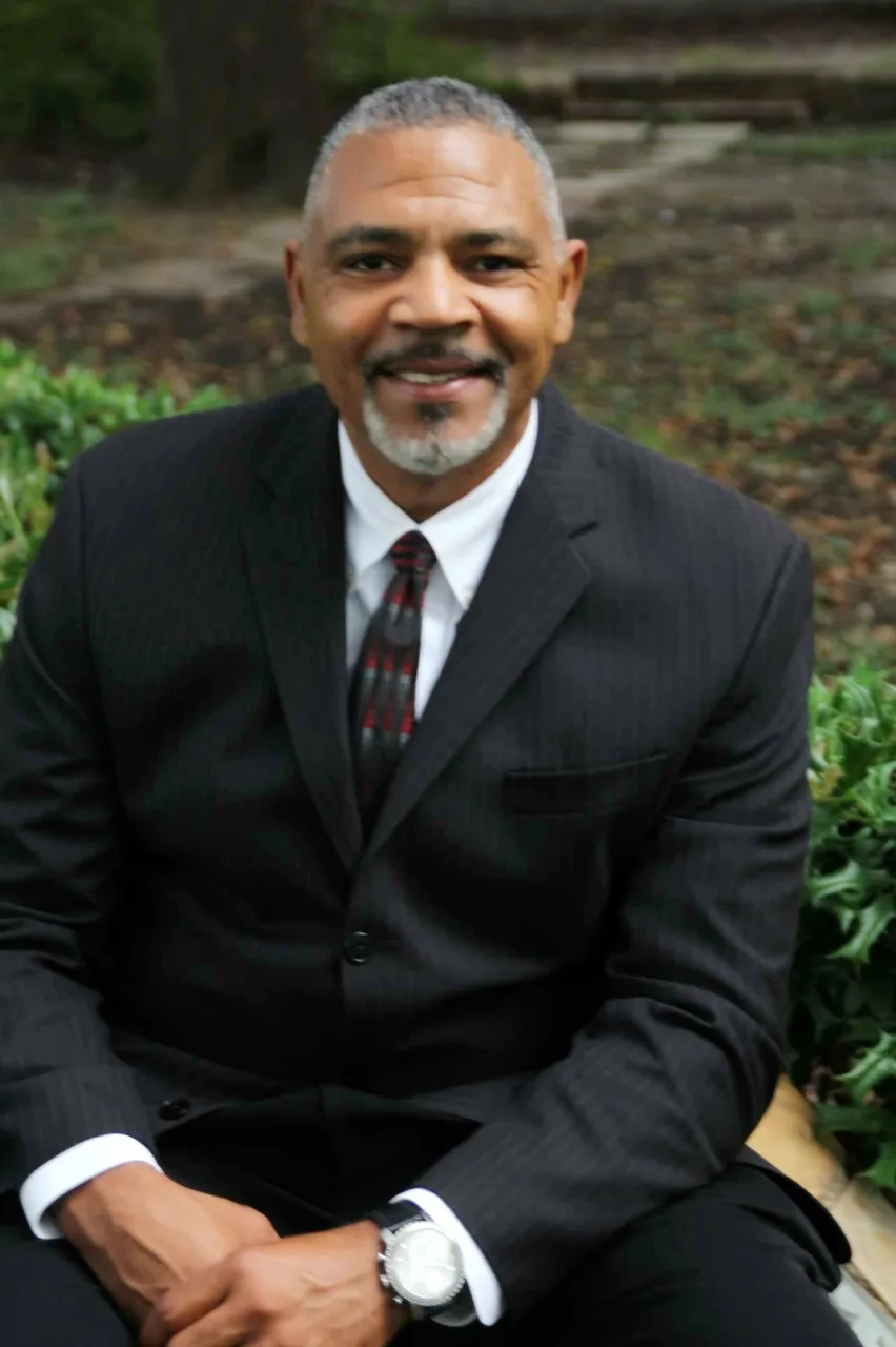 Derick Muhammad, seated outdoors in a dark suit.