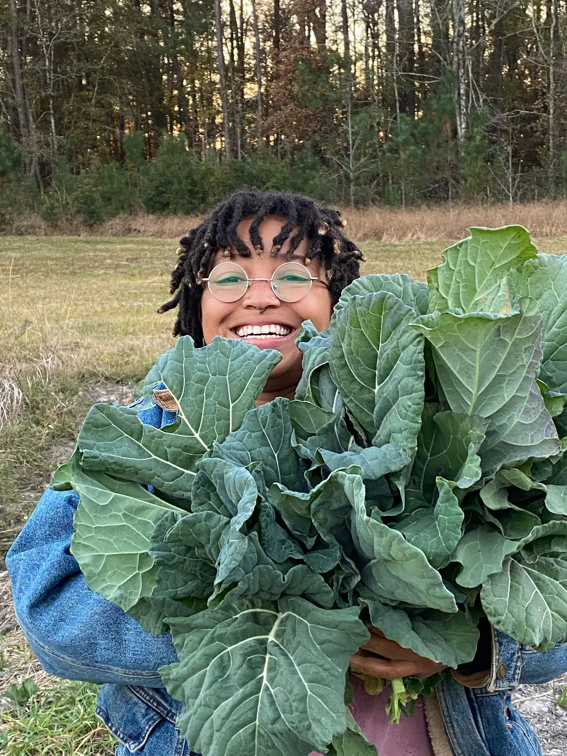 Sadia Pollard smiles behind an armful of freshly cut collard greens at the edge of a field.