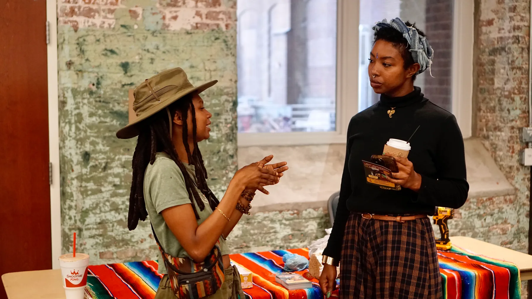 Two people talk beside a market table covered with a woven cloth.