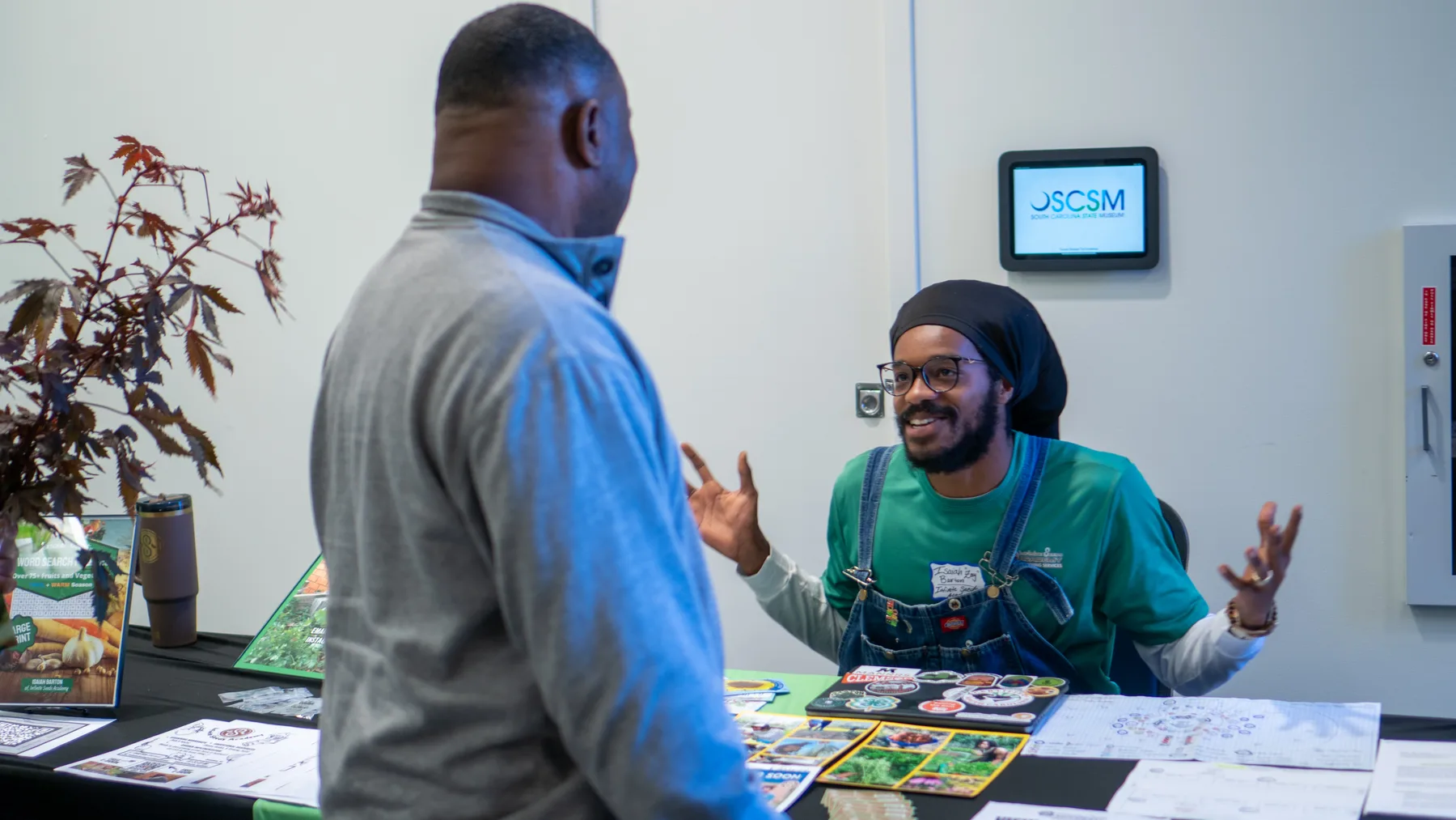 A person gestures while talking with a visitor beside a table of printed materials.