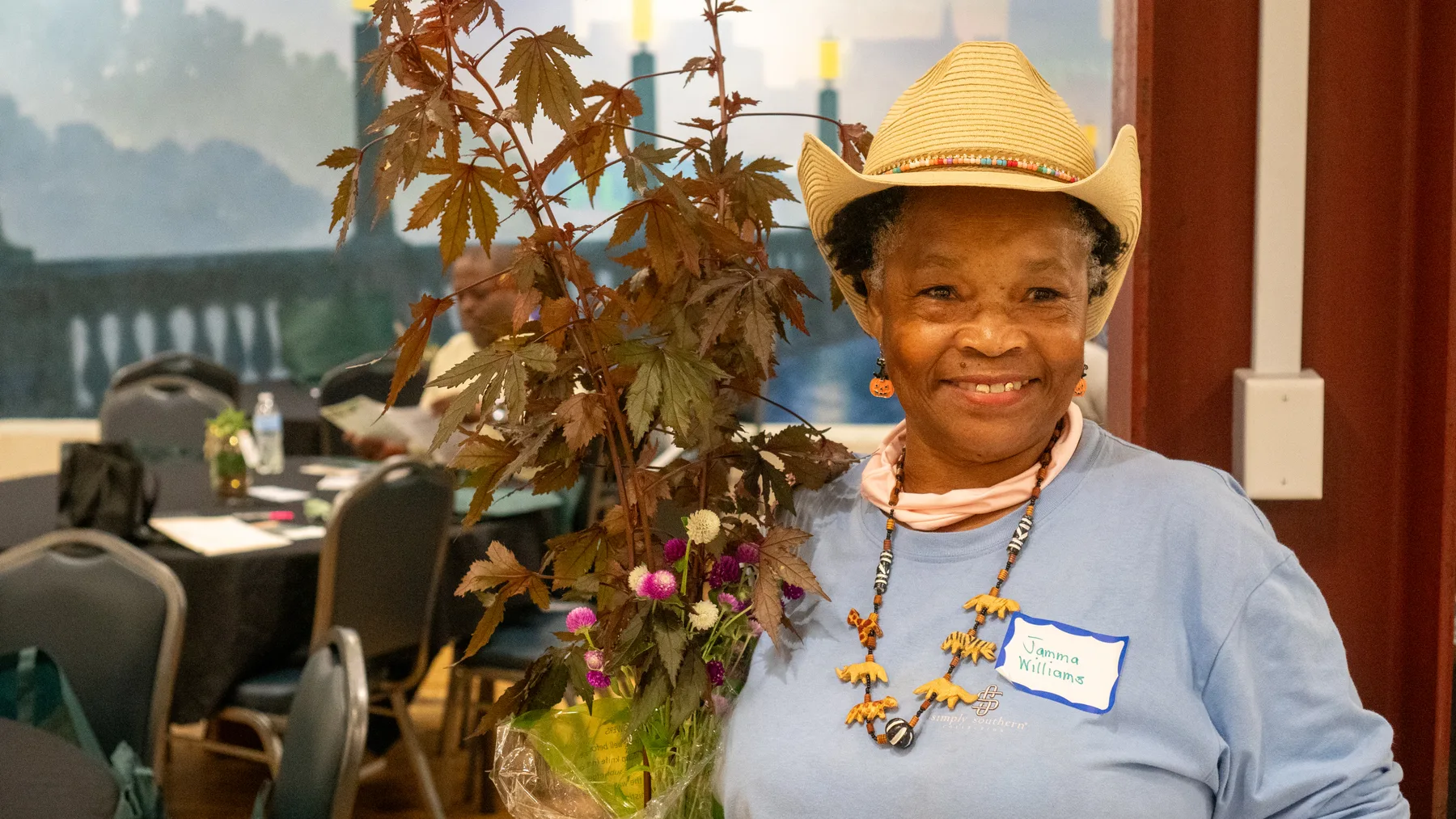 A person in a straw hat smiles beside an arrangement of plants.