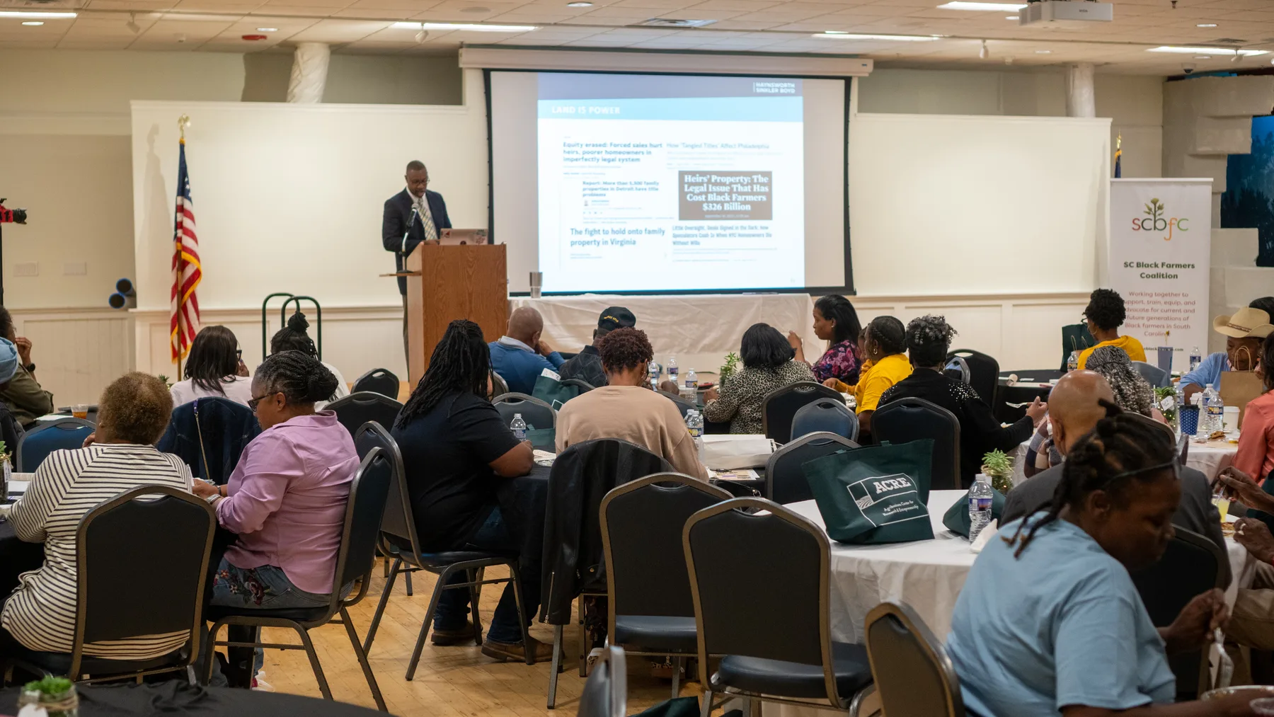 People fill a banquet room for a presentation on heirs’ property, with a South Carolina Black Farmers Coalition banner beside the screen.