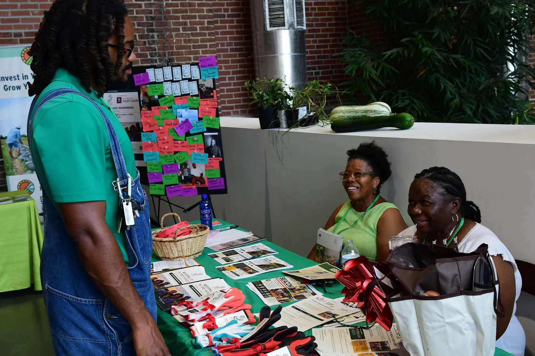 A man in overalls talks with two smiling women seated behind a table of printed materials.