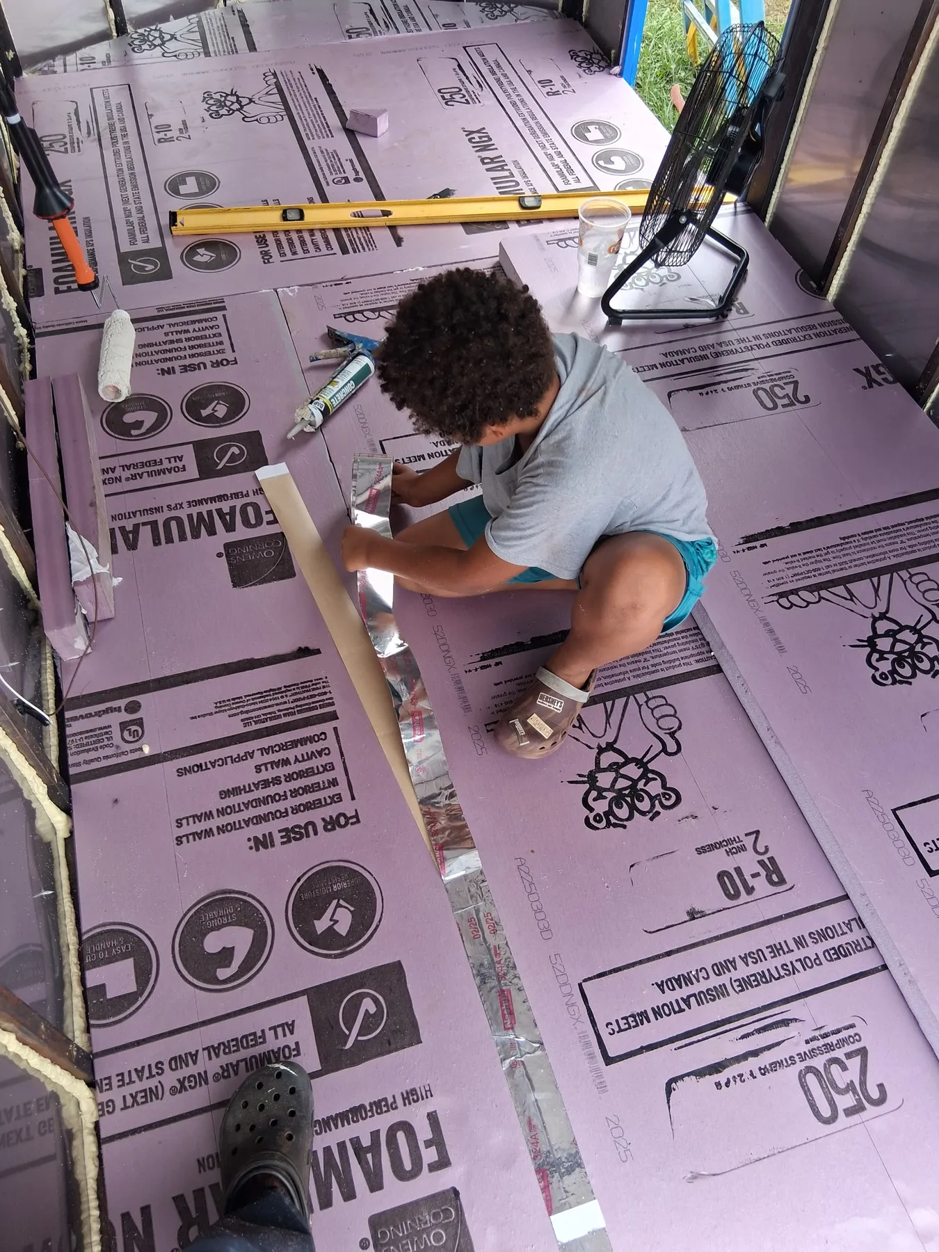 A person kneels on foam insulation boards inside the trailer, measuring a strip of wood.