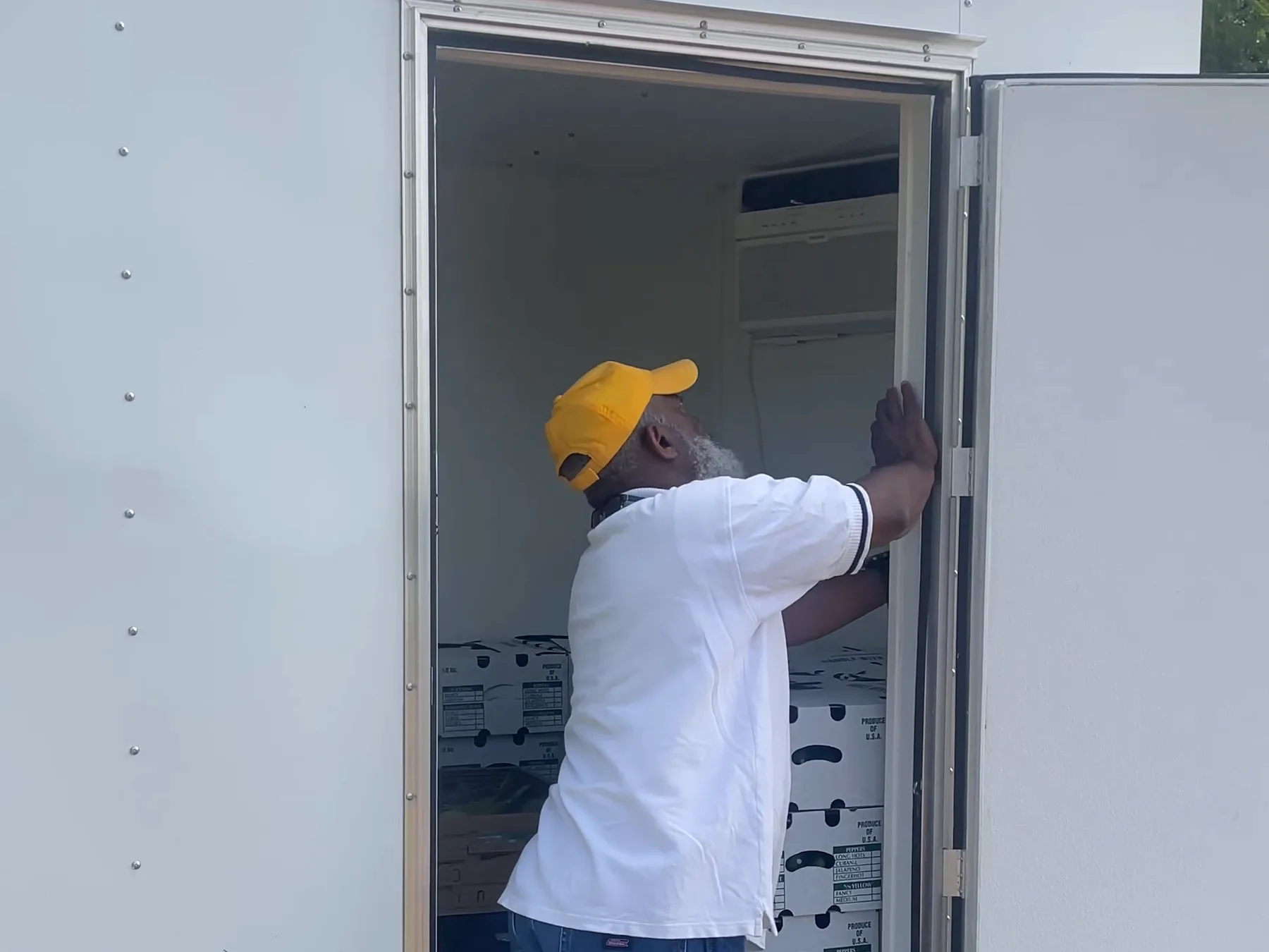A farmer in a yellow cap reaches into the open door of a white cold-storage trailer stacked with boxes.