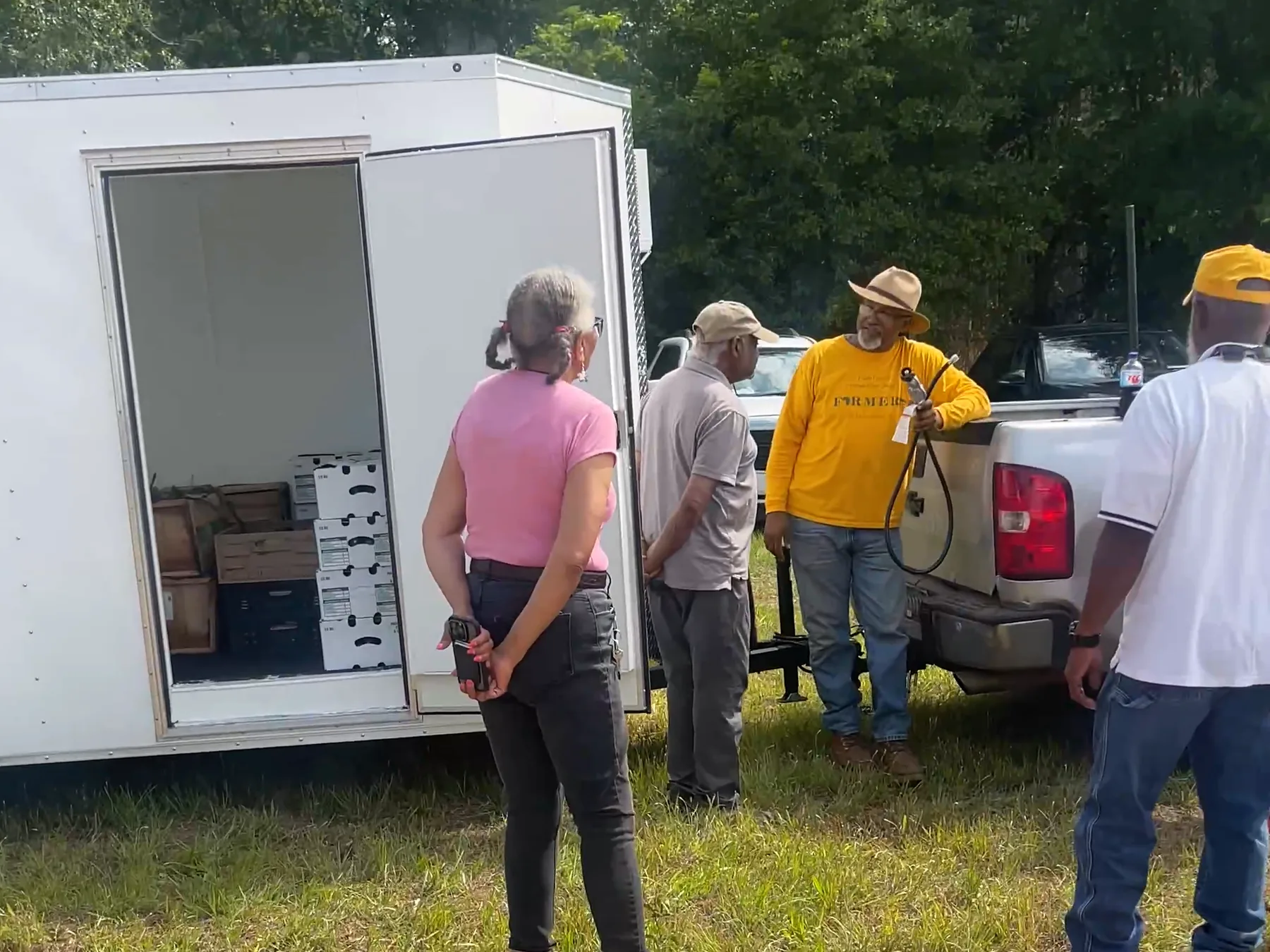 A group gathers beside one of the two mobile cold-storage units, its door open and stacked with produce boxes, at the edge of a farm field.