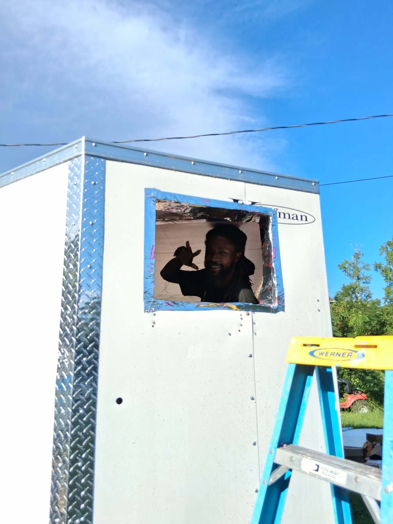 A person waves through the small window of a white trailer-mounted cold-storage unit under a blue sky.