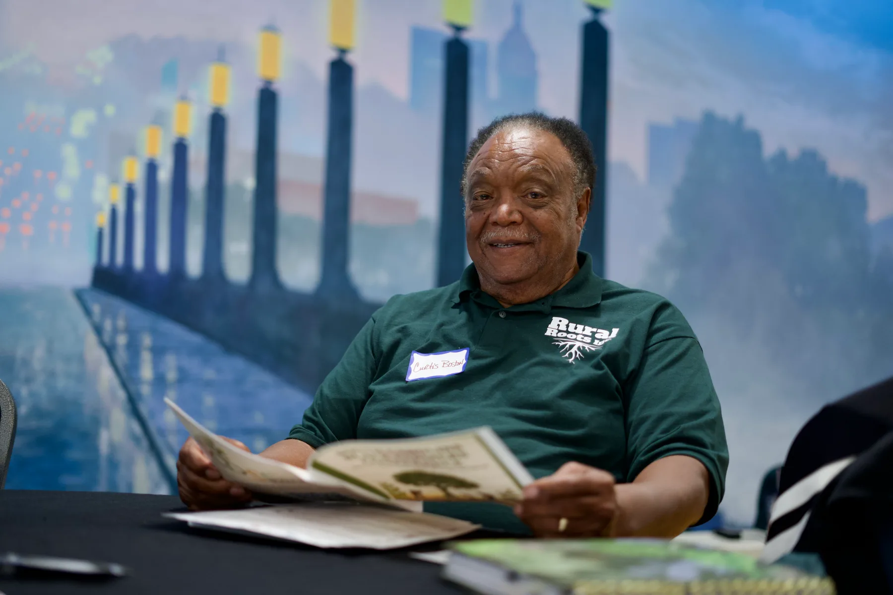 A person reads a printed publication in front of a painted landscape mural.