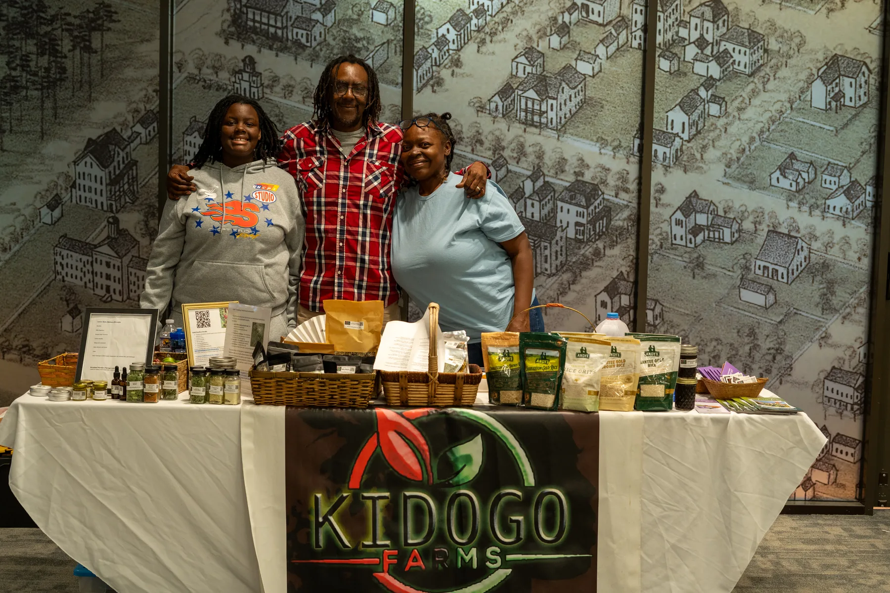 Three people stand behind a table of farm products and preserves.